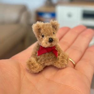 Puede incluir: Un oso de peluche en miniatura hecho de material de limpiapipas marrón difuso, con una pequeña nariz negra, ojos oscuros y una cinta roja brillante atada en un lazo alrededor del cuello. El oso está en posición sentada con pelaje texturizado.