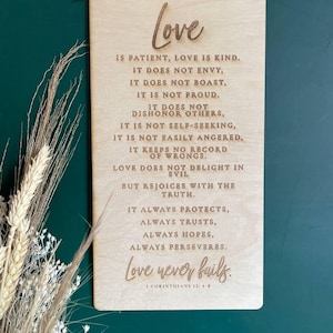 May include: A wooden sign with the text "Love" in a cursive font. The sign features a quote about love from the Bible, 1 Corinthians 13:4-8. The quote reads: "Love is patient, love is kind. It does not envy, it does not boast, it is not proud. It does not dishonour others, it is not self-seeking, it is not easily angered, it keeps no record of wrongs. Love does not delight in evil but rejoices with the truth. It always protects, always trusts, always hopes, always perseveres. Love never fails."