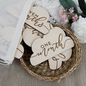 May include: A set of wooden cloud-shaped baby milestone cards with the words "one month", "seven months", and "six months" written on them. The cards are in a wicker basket and are surrounded by white fabric and flowers.
