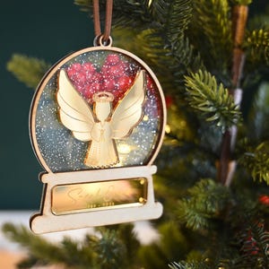 May include: A wooden Christmas ornament shaped like a snow globe, featuring a gold angel and red accents. The ornament has a small gold plaque with the name "Sarah E." and dates. It hangs from a brown cord and is set against a backdrop of a Christmas tree.