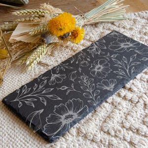 Puede incluir: Una bandeja de servir de pizarra negra con un diseño floral blanco. La bandeja está sobre una superficie de crochet de color crema.