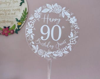 Könnte beinhalten: Transparenter Acryl-Kuchenaufsatz mit weißem Blumengirlanden-Design und dem Text "Happy 90th Birthday Janet". Der Aufsatz ist für einen Kuchen gedacht und eignet sich für einen 90. Geburtstag.
