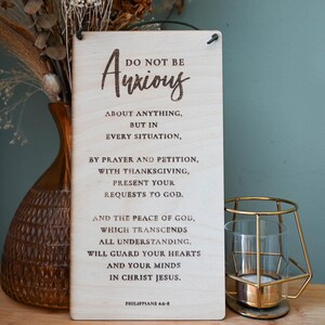 May include: Wooden sign with a white background and brown text. The sign reads "Do not be anxious about anything, but in every situation, by prayer and petition, with thanksgiving, present your requests to God. And the peace of God, which transcends all understanding, will guard your hearts and your minds in Christ Jesus. Philippians 4:6-7".