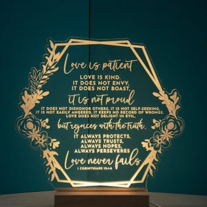 May include: A gold-coloured acrylic light-up sign with a floral design and the text "Love is patient. Love is kind. It does not envy, it does not boast, it is not proud. It does not dishonor others, it is not self-seeking. It is not easily angered, it keeps no record of wrongs. Love does not delight in evil, but rejoices with the truth. It always protects, always trusts, always hopes, always perseveres. Love never fails. 1 Corinthians 13:4-8."