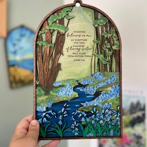 May include: Layered wooden artwork depicting a forest scene with a river and blue flowers. The piece includes the text "WHOEVER believes in me...rivers of living water...JOHN 7:38". It has a dark brown frame and a hole for hanging, suitable for wall display.