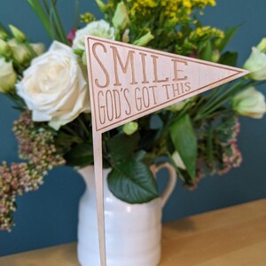 Puede incluir: Una bandera de madera con el texto "SMILE GOD'S GOT THIS" en un jarrón blanco con flores.