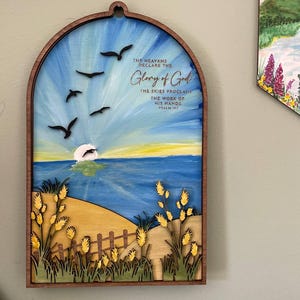 May include: Wooden wall art depicting a sunset over the sea with birds in silhouette. The artwork includes the text "THE HEAVENS DECLARE THE GLORY OF GOD" and "THE SKIES PROCLAIM THE WORK OF HIS HANDS PSALM 19:1". The frame is a dark brown colour.