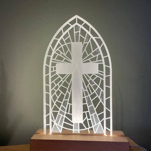 May include: A white stained glass cross lamp with a wooden base. The cross is in the shape of a stained glass window with a geometric pattern.