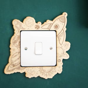 May include: A light switch cover with a wood veneer design featuring a rocket, planets, and stars. The design is in black and white.