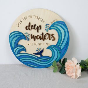 May include: A round wooden sign with a blue and green ocean wave design. The sign features a whale tail and the text "WHEN YOU GO THROUGH deep waters I WILL BE WITH YOU" and "ISAIAH 43:2".