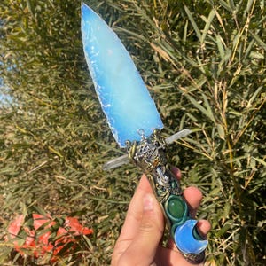 Puede incluir: Una daga de cristal azul y verde con un diseño de luna creciente. La daga tiene un mango de metal con un diseño floral y un amuleto de cristal.