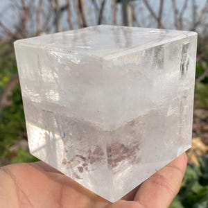 Puede incluir: Un gran cristal cúbico transparente con facetas internas visibles. El cristal es liso y pulido, con un ligero brillo.