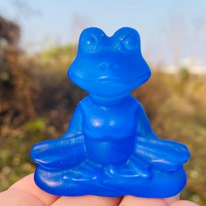 Puede incluir: Una figurita de rana de vidrio azul sentada en una postura meditativa. La rana tiene un acabado liso y brillante y es de un tono azul vibrante.