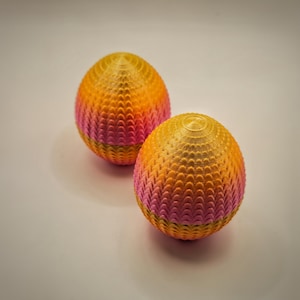 Könnte beinhalten: Zwei dekorative, eiförmige Objekte mit strukturierter Oberfläche. Die Eier wechseln die Farbe von Gelb über Orange zu Rosa. Die Objekte sind wahrscheinlich 3D-gedruckt und vor einem neutralen Hintergrund platziert.