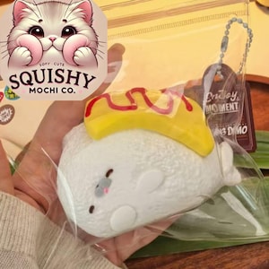 Può includere: Un giocattolo squishy bianco a forma di foca con una copertura gialla e rossa, confezionato in un sacchetto di plastica trasparente. La borsa ha un'etichetta con la scritta "Enjoy Moment". L'immagine mostra anche un logo "Squishy Mochi Co." con un simpatico gatto.