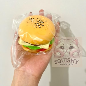 Op de afbeelding: Een squishy speelgoed in de vorm van een hamburger, met een gele broodje bestrooid met zwarte en witte sesamzaadjes. De burger bevat een rode plak tomaat, groene sla en een gele plak kaas. Het speelgoed zit in een doorzichtige plastic verpakking. Een sticker met een kattenillustratie en de tekst "Squishy Mochi Co." is zichtbaar.