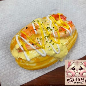 May include: A glazed, golden-yellow mochi donut topped with shrimp, yellow sauce, white drizzle, and black sesame seeds. The donut sits on a white paper. A logo with a cute cat face and the text "Squishy Mochi Co." is in the bottom right corner.