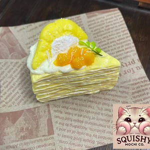Op de afbeelding: Een gele crêpe taartpunt met een mangotopping en room, gepresenteerd op een achtergrond in krantenstijl. De afbeelding bevat een logo met de tekst "Squishy Mochi Co."