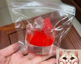 Könnte beinhalten: Eine transparente Plastiktüte mit einem kleinen, durchsichtigen, quadratischen Behälter, der mit roter Flüssigkeit, Eiswürfeln und einem roten erdbeerförmigen Objekt gefüllt ist. Ein Cartoon-Kätzchen mit dem Wort "SQUISHY" befindet sich in der unteren rechten Ecke.