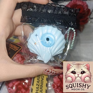 May include: A light blue seashell-shaped charm with a central eye design and small star accents, enclosed in a clear plastic pouch featuring a black ruffled lace trim at the top and a light green beaded chain attached to the side.