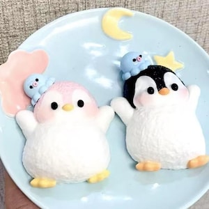 May include: Two white penguin figurines with pink and black accents. Each penguin has a smaller blue octopus figurine on top of its head. The penguins are on a light blue plate with a yellow crescent moon and a yellow star.