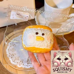 Könnte beinhalten: Ein weiß-gelbes Mochi-Dessert in Entenform in einer transparenten Plastiktüte. Das Mochi hat einen gelben Schnabel und braune Augen. Die Tüte ist mit einem dekorativen Rand verschlossen. Das Logo "Squishy Mochi Co." ist sichtbar.