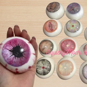 Könnte beinhalten: Eine Sammlung von 12 quietschigen Mochi in Augenform, jede mit einer anderen Farbe und einem anderen Design. Die Mochi sind aus einem weichen, quietschigen Material gefertigt und perfekt zum Drücken und Spielen. Die Mochi sind in verschiedenen Farben erhältlich, darunter Pink, Lila, Blau, Grün und Gelb. Jedes Mochi hat ein anderes Design, darunter eine schwarze Pupille, eine weiße Sklera und eine Vielzahl von farbigen Iris.