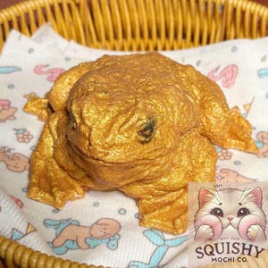 Puede incluir: Un juguete blando en forma de rana dorada. El juguete está hecho de un material suave y blando y tiene un diseño de rana realista. El juguete está sentado sobre una tela con estampado blanco y azul.