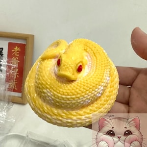 Puede incluir: Un juguete blando de color amarillo y blanco con forma de serpiente y ojos rojos. El juguete es suave y tiene una superficie texturizada. El juguete está siendo sostenido en una mano.