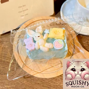 May include: A blue, square mochi dessert decorated with a bunny, teddy bear, star, and strawberry. The mochi is in clear plastic on a wooden plate. The image also shows a logo that says "Squishy Mochi Co."