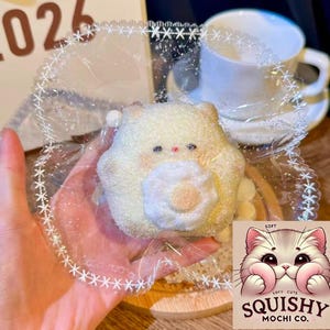 May include: A soft, yellow mochi dessert shaped like a bear, with a white flower-like center, wrapped in clear plastic. The image includes a white teacup and saucer, and a logo that says "Squishy Mochi Co."