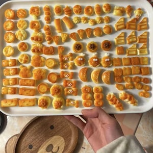 Può includere: Un vassoio bianco pieno di piccole miniature di cibo in resina gialla e arancione. Le miniature sono disposte in file e includono varie forme, come ciambelle, pane e biscotti.