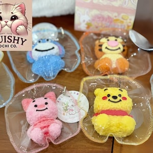 Op de afbeelding: Een assortiment kleurrijke mochi-lekkernijen in de vorm van stripfiguren, waaronder een blauw personage, een tijger, een roze varken en een gele beer. Elke mochi staat op een helder, bladvormig schaaltje. Het logo van "Squishy Mochi Co." is zichtbaar.