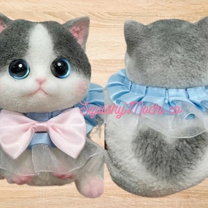 Puede incluir: Un gato de peluche gris y blanco con ojos azules que lleva un collar de volantes azul claro con un lazo rosa. El gato está sentado sobre una superficie de madera.