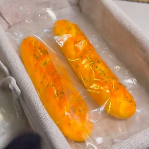 Puede incluir: Dos panes amarillos y naranjas están en bolsas de plástico transparentes. El pan está en una cesta de tela blanca.