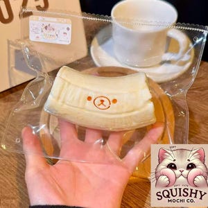 May include: A packaged, cream-colored dessert with a bear face design, held in a clear plastic bag. The dessert sits on a wooden plate, with a white teacup and saucer in the background. The image also includes a logo that says "Squishy Mochi Co."
