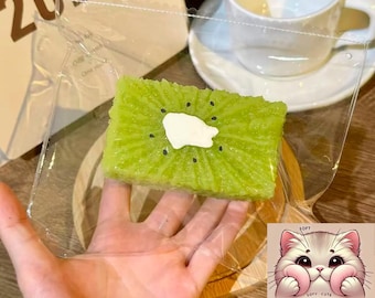 Könnte beinhalten: Ein grünes Mochi-Dessert in Kiwi-Form mit weißer Sahnehaube, gehalten in einer transparenten Plastikverpackung. Das Mochi liegt auf einem Holzbrett, im Hintergrund eine Tasse und Untertasse. Das Logo von Squishy Mochi Co. befindet sich unten rechts.