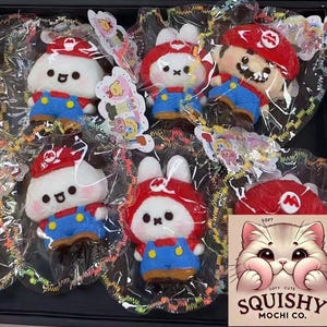 Op de afbeelding: Een verzameling zachte, squishy mochi-lekkernijen in de vorm van stripfiguren, waaronder een konijn en een personage met een rode hoed en blauwe overalls. Elke mochi is afzonderlijk verpakt in doorzichtig plastic. De afbeelding bevat een logo met een kat.
