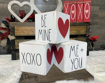 May include: Four white wooden blocks stacked on top of each other. The blocks have different phrases written on them: "Be Mine", "XOXO", "Me + You", and a heart. The blocks are decorated with red hearts.