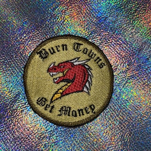 May include: A round, tan embroidered patch with a red dragon and the text "Burn Towns Get Money".