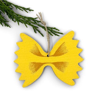 May include: A yellow wooden ornament shaped like farfalle pasta, with a brown, textured design in the center. A twine loop is attached for hanging. Green sprigs are in the upper left corner, suggesting a holiday theme.