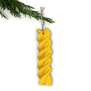 May include: A yellow wooden ornament in the shape of a twisted pasta noodle, suspended from a twine string. The ornament hangs from a sprig of green foliage against a white backdrop. The pasta-shaped decoration has a textured surface.