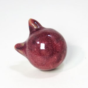 Può includere: Una piccola statuetta di ceramica di un gatto con una glassa rosa. Il gatto ha un corpo rotondo e una piccola coda appuntita.