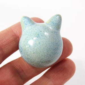 May include: A small ceramic cat figurine with a light blue and white speckled glaze. The cat has a rounded body and two small ears.