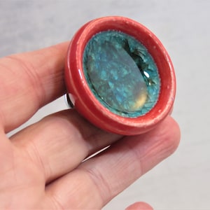 Puede incluir: Un pomo redondo de cerámica con un borde rojo y un centro azul verdoso. El centro tiene un acabado texturizado e iridiscente.