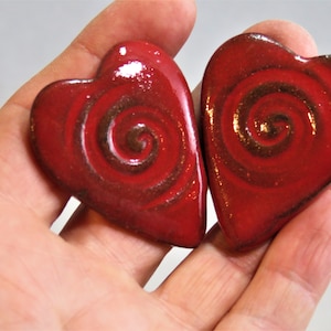 May include: Two red ceramic hearts with a swirling gold design. The hearts are held in a hand.