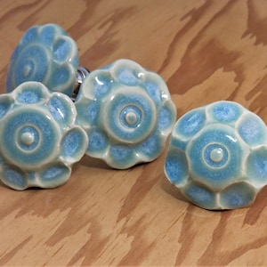May include: Four ceramic cabinet knobs in a light blue and white glaze with a flower design. The knobs have a raised center circle and a scalloped edge.