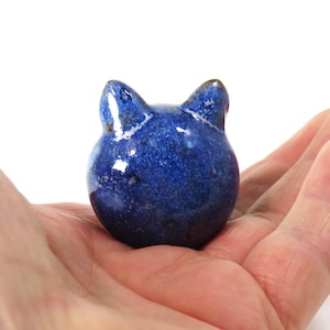 May include: A small, blue ceramic cat figurine with a speckled glaze. The cat is sitting on a hand, facing away from the camera.