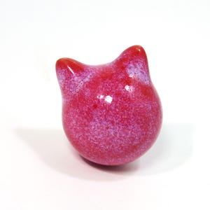 May include: A small, pink ceramic cat figurine with a speckled glaze. The cat is sitting upright with its ears perked up.