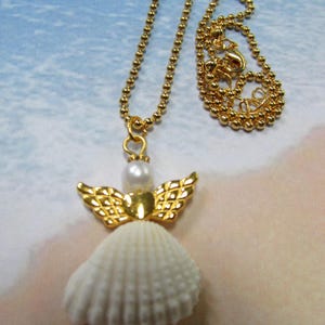 May include: A gold necklace with a seashell angel pendant. The pendant features a white seashell body, gold wings, and a pearl head. The necklace chain is made of small gold beads, and the clasp is visible.